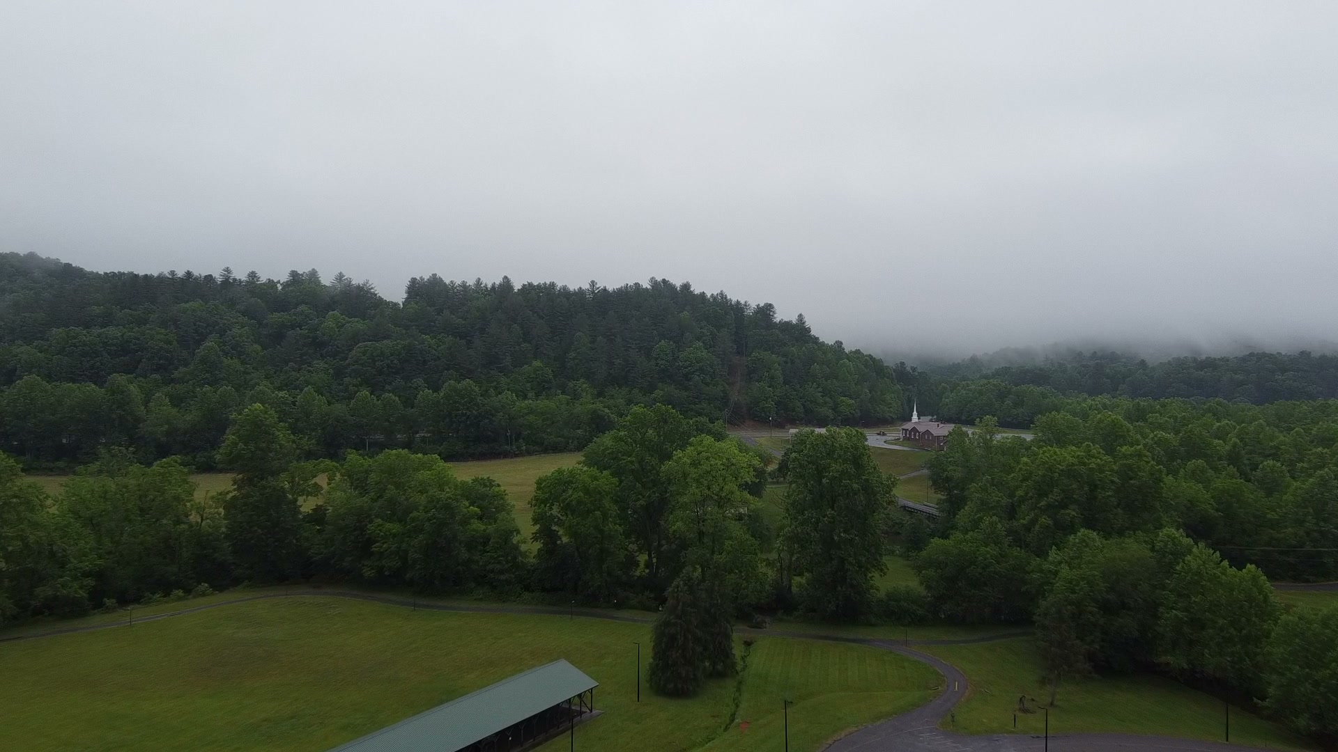 An aerial view over Parker Meadows: flat open fields in the foreground, a wooded hillside rising into low cloud behind. The flat ground in front is where the first flight worked. The rising woods in back are where the second one didn't.